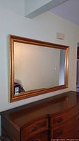 Full view of the rectangular mirror with gold frame, mounted on wall above a wooden chest of drawers.