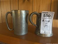 Two pewter vessels, a cylindrical pint mug and a jug with handle and base rings, shown side by side on wood surface.