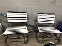 Two white slatted outdoor chairs with coated metal frames side by side against a wall.