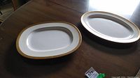 Two rectangular white platters with intricate gold patterned trimming, displayed on wooden table