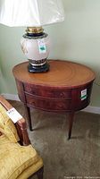 View of oval wood side table with reddish finish, leather inlay top with tooling, lamp base on top for scale.