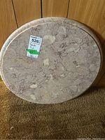 Round stone table top leaning against wood wall, showing marbled light beige and cream colors with a glossy finish.