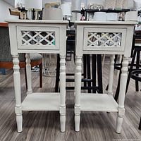 Front view of two matching off-white decorative accent tables with mirrored drawers and turned legs on a hardwood floor.