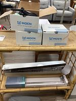 Three Moen boxes stacked with one open showing a black shower fixture. Below, two towel bars packaged in rectangular boxes, one from Kohler and one from AquaSource.