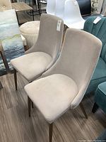 Two matching armless chairs with light beige micro suede upholstery and wooden legs displayed on a wooden floor, showing the front and side views.