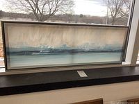 Framed abstract landscape canvas print positioned on a windowsill. Horizontal rectangular frame measuring approximately 37x13 inches. The painting has soft blues, beige, and gray hues.