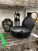 Photo of the full set composed of one black resin sculpture, one black ceramic pitcher, one decorative bowl, and one floral vase on a metal rack.