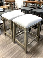 Photo showing a pair of wood framed stools with light beige fabric upholstered square seats, positioned on a laminated wood floor.