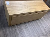 Angled front view of storage bench showing overall shape, light wood color, and the crease for the slide-out drawer front.