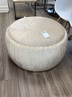 Front and angled view of round beige velour upholstered ottoman on wood floor.