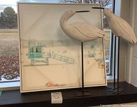 Pair of resin stork figures on black metal legs placed next to framed 18x18 canvas print of beach scene on window ledge.