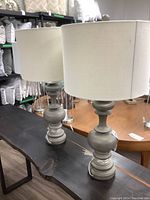 Pair of gray resin table lamps with fabric drum shades placed on a black bench with a wood grain top, displayed inside a room with other cushions and lamps.