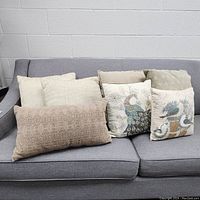 Photo showing assorted throw cushions on a gray sofa including beige woven cushions, printed peacock and bird design cushions, and a long taupe textured cushion.