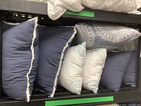 Seven assorted throw cushions arranged on a shelf showing blue cushions with white fringed edges, white cushions with blue embroidery, and a grey patterned cushion.