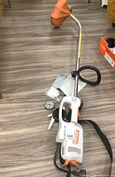 Gesamtes Trimmergerät mit oranger Schutzvorrichtung, schwarzem Tragegurt und Zubehör auf dem Holzfußboden.