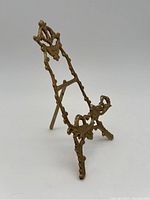 Front angle view of a brass decorative stand highlighting the curved easel back and the intricate design of the base.