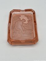 Top view of pink glass ashtray showing seated nude relief design embedded in the base, surrounded by beveled edges and cigarette notches.