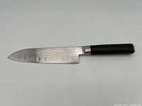 Full length view of the Shun Damascus steel Santoku knife showing blade and dark handle with metal collar, laid horizontally on white background.