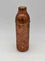 Solid copper water bottle standing upright with visible tarnish and patina on surface, domed copper screw cap closed.