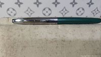 Fountain pen showing full length of the closed pen, green barrel and silver cap visible, placed on cloth surface with designer pattern background.