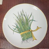 Top view of a white round plate featuring a botanical illustration of chives tied with a yellow string with gold rim.