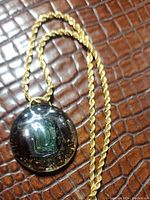 Front view of the round pendant with green letter D and gold fleck design on gold-toned rope chain, displayed on brown textured surface.