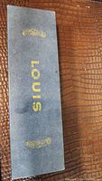 Closed blue leatherette watch box with gold Louis logo and design on top, placed on brown textured surface.