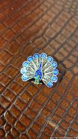 Close-up photo of small peacock brooch on textured brown background, showing blue and green enamel on silver filigree body and feathers.