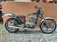 Side view of Suzuki GS400 motorcycle frame with engine block mounted, no seat or fuel tank, black frame, chrome fenders on wheels.