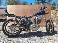Side view of black motorcycle frame with engine and rear wheel attached, displayed outdoors on gravel ground with wood board background