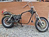 Side view photo showing the partial black frame, rear wheel with Firestone tire, rear suspension shocks, front forks, and high-rise handlebars without seat, engine, or fuel tank.