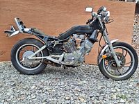 Side view of 1984 Yamaha XV500 Virago motorcycle with frame, engine, wheels and seat frame visible, placed on gravel ground with wooden backdrop.