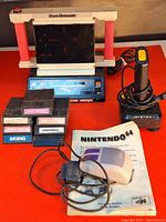 Photo of Atari 2600 Cosmic Commander console with six Atari 2600 game cartridges in front, Nintendo 64 mouse on instruction booklet, and joystick with base and coiled cord on side.
