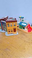 Photo showing two vintage Fisher Price Sesame Street buildings on a wooden surface: a yellow multi-story building resembling Mr Hooper's Store and a smaller white and green building with a red spiral slide.