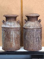 Front view of two rusty metal milk cans with lids and handles placed side by side with a ruler behind them showing approximate height.