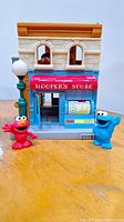 Front view of Mr Hoopers Store playset with blue awning, two Sesame Street figurines (Elmo and Cookie Monster) positioned in front, and an additional figure visible through upper window.