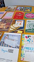 Overview of multiple mid-century pamphlets and recipe books spread on table; includes colorful covers and various titles.