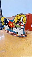Side view of vintage toddler rocking horse showing litho print design with yellow mane, red saddle featuring maple leaf, and striped rocker base.
