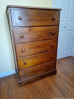 Front view of a medium brown wooden dresser with five drawers, all with round wooden knobs. There is one knob with noticeable surface damage.