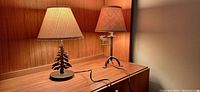 Image of two rustic cabin style table lamps; one with a metal tree motif and round base, the other with a metal moose figure and tripod base. Both have beige textured shades and cords extended on wooden cabinet surface.