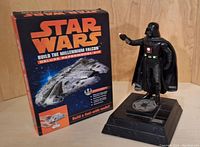 Darth Vader action figure on standing platform with Imperial logo next to unopened Millennium Falcon paper model kit box