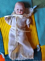 Front view of porcelain baby doll dressed in white gown with lace trim, showing head, arms, and legs made of porcelain, lying on green and yellow fabric.