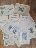 Photo showing multiple aged newspaper pages spread out on a wooden surface. Visible are various headlines, articles, and partial financial statements.