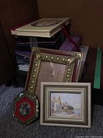Stacked photo albums in various colors and pile of assorted picture frames including one with a coastal town painting and a larger gold-toned frame