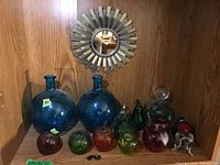 Group photo of art glass collection consisting of two large blue bottles and several small colorful glass apples and spheres arranged under a sunburst mirror.