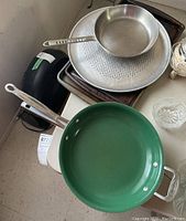 Green 12-inch skillet with metal handle on top of stack including other skillets, perforated pizza pan and cookie sheets