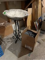 White resin birdbath with floral design on black metal stand, positioned on concrete floor near wall with cardboard boxes and vinyl records nearby.