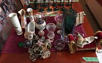 Wide view of table with various decorative figurines, glass items, and vases arranged on a red tablecloth, including shoe and boot figurines, glass ship in bottle, glass bowl, and paperweights.