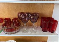 Shelf with coloured glassware including purple wine glasses, ruby red drinking glasses, clear large bowl, and ruby red small bowls and dishes.