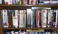 Shelf of assorted DVD movie cases showing various genres including comedy, kids movies, drama, action, and Hollywood classics arranged vertically on a wooden shelf